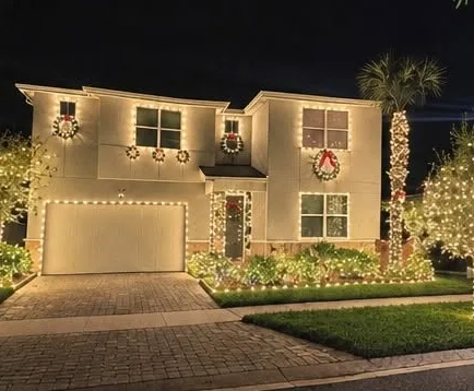 Two-story home with warm white roofline lights, wreaths on every window and a wrapped palm tree